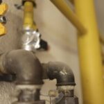 Close-up of a gas pipe with a yellow valve handle and threaded fittings mounted on a textured wall, with more piping in the background.