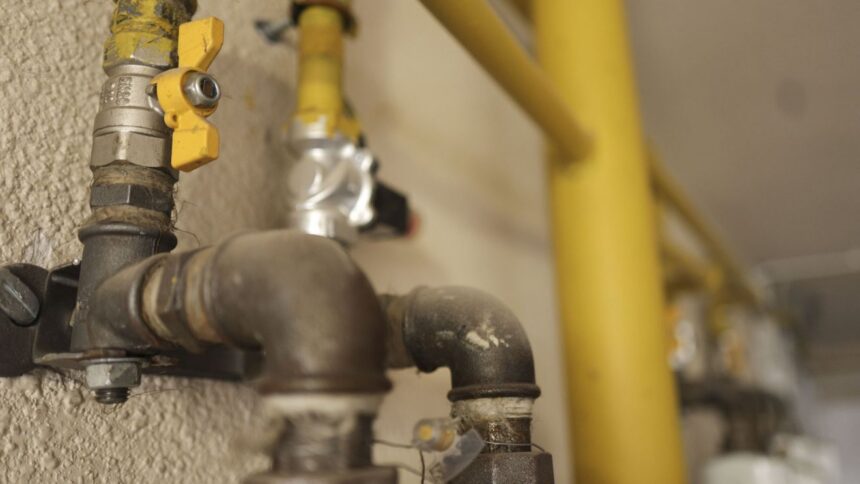 Close-up of a gas pipe with a yellow valve handle and threaded fittings mounted on a textured wall, with more piping in the background.