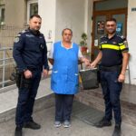 Two police officers hand a dark bag to a woman in a blue vest outside a building entrance.