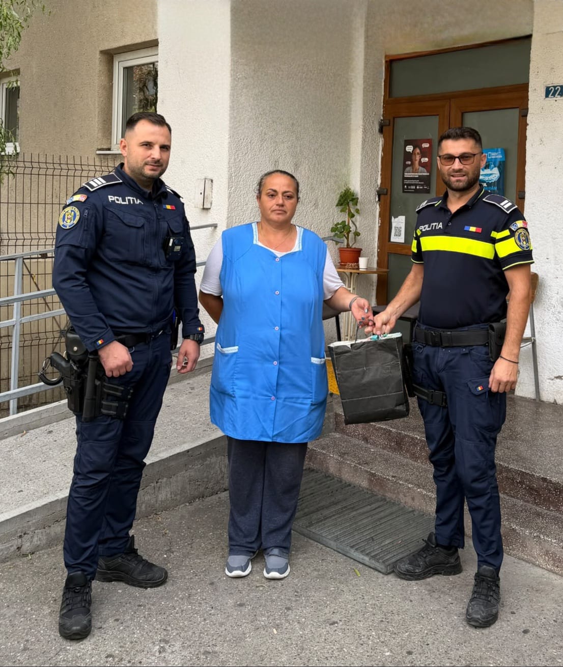 Two police officers hand a dark bag to a woman in a blue vest outside a building entrance.