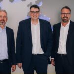 Three businessmen in dark suits stand side by side in front of a world-map wall in an office setting.