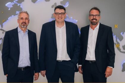 Three businessmen in dark suits stand side by side in front of a world-map wall in an office setting.