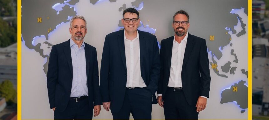 Three businessmen in dark suits stand side by side in front of a world-map wall in an office setting.
