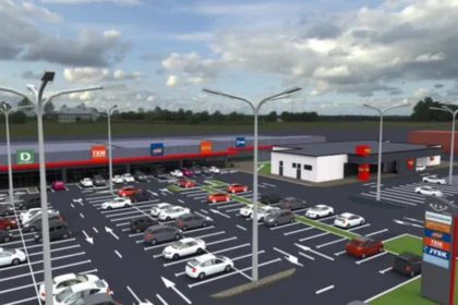 Large daytime parking lot with numerous cars in front of a shopping center and a small white building at the center-right.