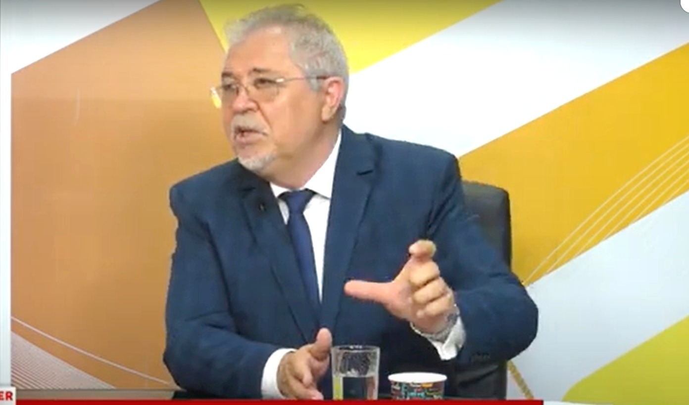 Man in a dark suit and tie speaks during a televised interview, gesturing with his right hand; glass and cup on the desk in front of him.