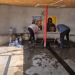 Two men filling water from a trough inside a plain room, with religious icons on the wall and rows of bottled water along the benches.
