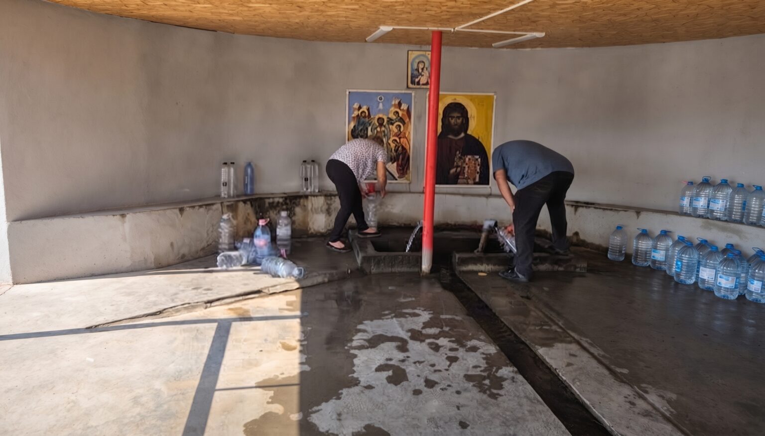 Two men filling water from a trough inside a plain room, with religious icons on the wall and rows of bottled water along the benches.