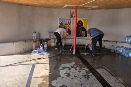Two men filling water from a trough inside a plain room, with religious icons on the wall and rows of bottled water along the benches.