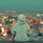 Person in a gray hoodie sits with back to the camera, overlooking a blurred city lights at dusk/nightfall from a high vantage point.
