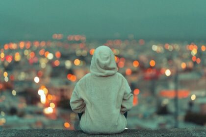 Person in a gray hoodie sits with back to the camera, overlooking a blurred city lights at dusk/nightfall from a high vantage point.