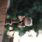 Wooden kendama toy hanging from a rope tied to a wooden post outdoors, with blurred green foliage in the background.