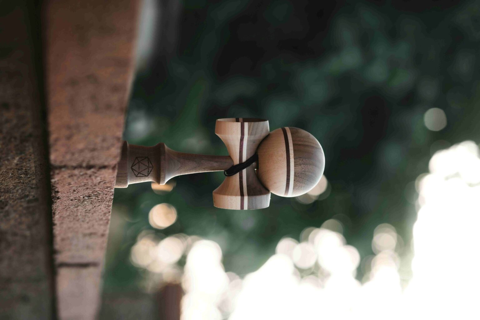 Wooden kendama toy hanging from a rope tied to a wooden post outdoors, with blurred green foliage in the background.