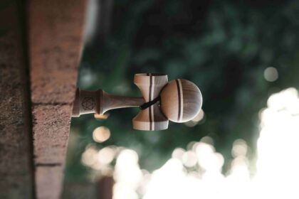 Wooden kendama toy hanging from a rope tied to a wooden post outdoors, with blurred green foliage in the background.