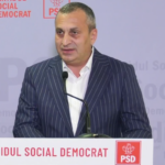 Man in a dark pinstripe suit speaks at a podium with a PSD banner and logo in the background.