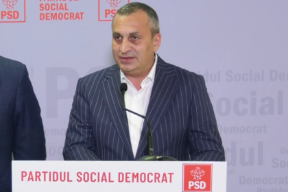 Man in a dark pinstripe suit speaks at a podium with a PSD banner and logo in the background.