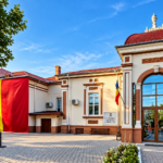Municipal building with a large vertical Romanian flag banner on the left, red-tiled roof, glass arched entrance, and clear blue sky.