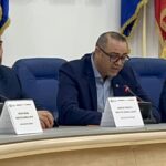 Three men in suits seated at a panel table with microphones and name placards, flags behind them in a formal conference setting.
