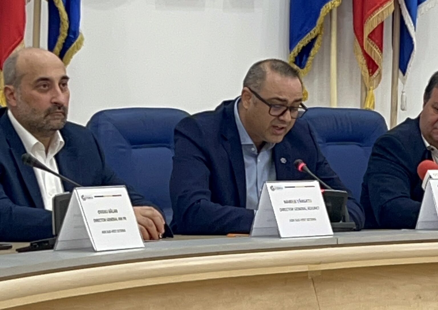 Three men in suits seated at a panel table with microphones and name placards, flags behind them in a formal conference setting.
