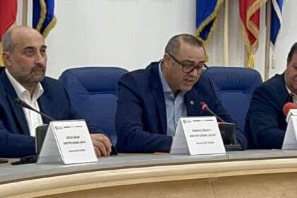 Three men in suits seated at a panel table with microphones and name placards, flags behind them in a formal conference setting.