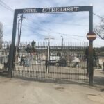Entrance to a cemetery with a tall wrought-iron gate reading 'Cimitirul Strehaer' and a visible row of grave markers beyond the gate.