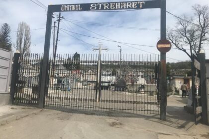 Entrance to a cemetery with a tall wrought-iron gate reading 'Cimitirul Strehaer' and a visible row of grave markers beyond the gate.