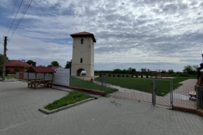Beige tower with a red-tiled pyramid roof in a fenced park under a cloudy sky.