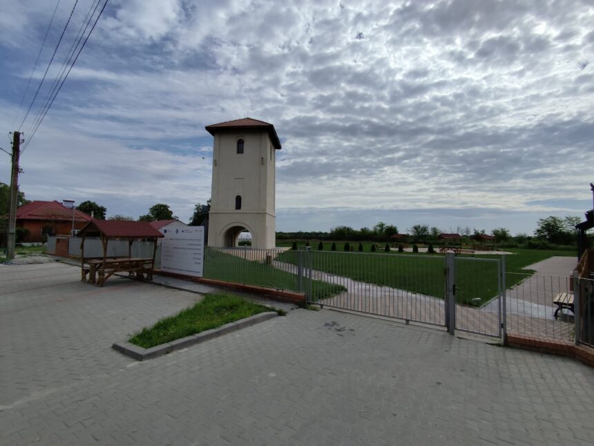 Beige tower with a red-tiled pyramid roof in a fenced park under a cloudy sky.