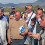 Group of men outdoors in a rural field, some wearing police caps, gathered around a man holding papers while others listen.