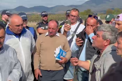 Group of men outdoors in a rural field, some wearing police caps, gathered around a man holding papers while others listen.