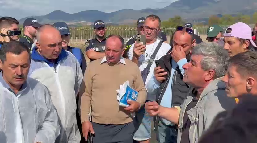 Group of men outdoors in a rural field, some wearing police caps, gathered around a man holding papers while others listen.