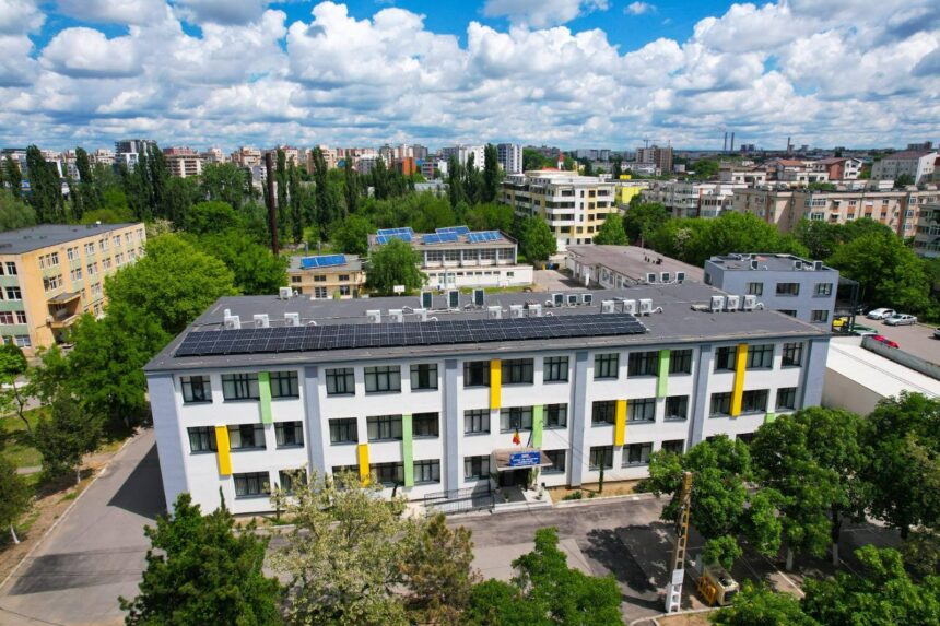 Aerial view of a modern white building with yellow and green vertical accents and solar panels on the roof, in a green urban area with other buildings nearby.