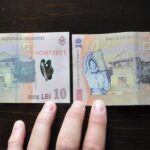 Two ten‑lei Romanian banknotes laid side by side on a dark surface, with a few fingers visible at the bottom.