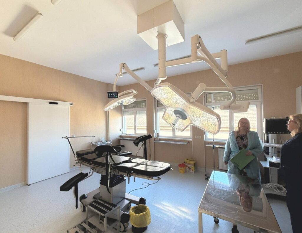 1000262375 - Jurnalul Olteniei – Știri din Craiova și Oltenia Operating room with a patient table and ceiling-mounted surgical lights; two medical staff in scrubs stand near a stainless table.