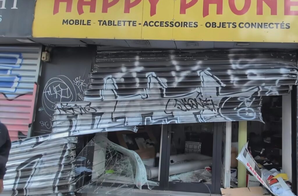 Shopfront with a yellow sign and red lettering; the metal shutter is severely damaged and covered in graffiti, with broken glass and debris visible inside the store.