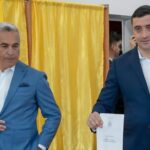 Two men in blue suits stand near a yellow curtain; the man on the right holds a document with a crest visible?