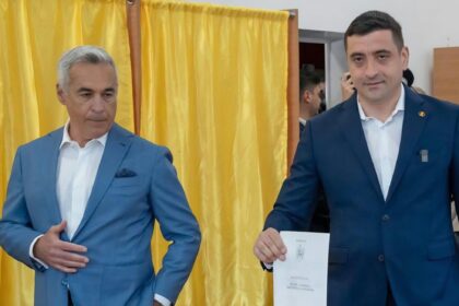 Two men in blue suits stand near a yellow curtain; the man on the right holds a document with a crest visible?