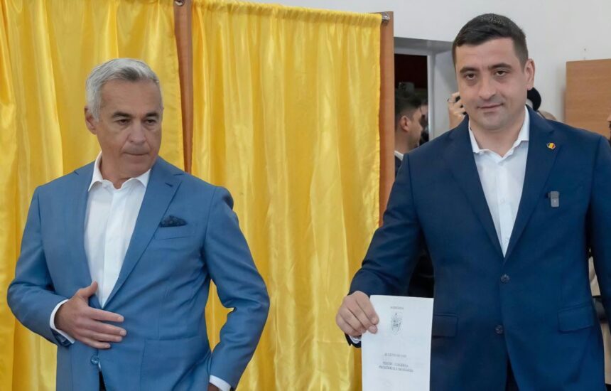Two men in blue suits stand near a yellow curtain; the man on the right holds a document with a crest visible?