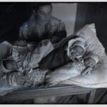 A woman sits beside a crib with three newborn babies wrapped in blankets on the bedding nearby.