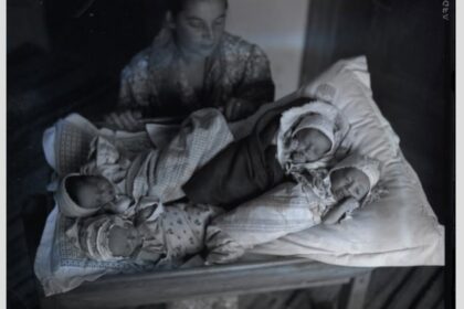 A woman sits beside a crib with three newborn babies wrapped in blankets on the bedding nearby.