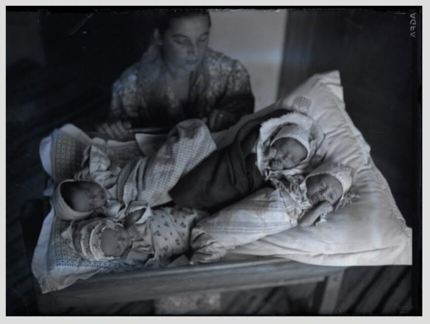 A woman sits beside a crib with three newborn babies wrapped in blankets on the bedding nearby.