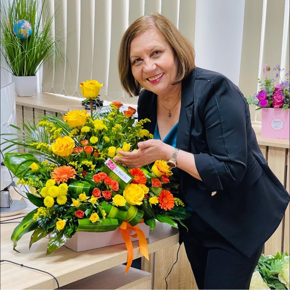 - Jurnalul Olteniei – Știri din Craiova și Oltenia Smiling woman in a dark blazer stands beside a large yellow and orange flower bouquet on a desk.