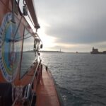 Orange rescue boat side view with equipment, sun reflecting on water, harbor and lighthouse in distance.