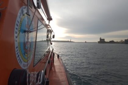 Orange rescue boat side view with equipment, sun reflecting on water, harbor and lighthouse in distance.