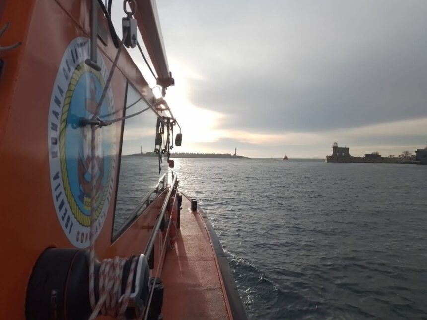 Orange rescue boat side view with equipment, sun reflecting on water, harbor and lighthouse in distance.