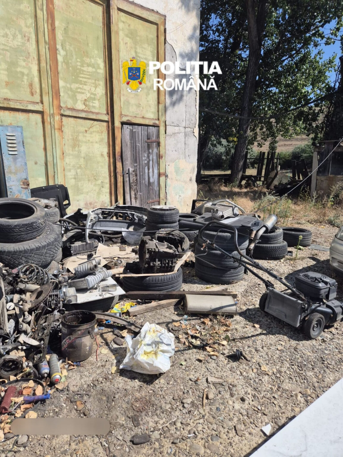 Outdoors, a junkyard scene with tires and car parts scattered on a dirt ground in front of a weathered building, with a Romanian Police logo overlay.