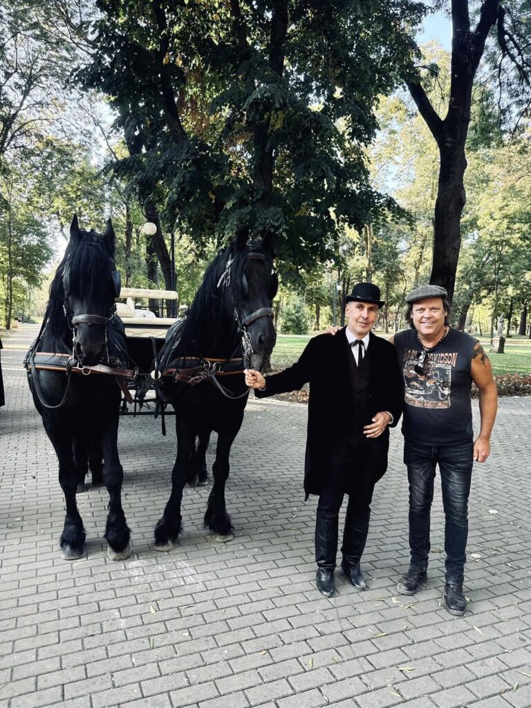 Two black horses stand side by side, hitched to a carriage, as two men pose in a park; one wears a suit and bowler hat, the other a gray cap and graphic t-shirt.