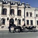 Horse-drawn carriage with a driver and two passengers in formal attire, pulled by a black horse in front of an ornate white stone building with arched windows and decorative details under a clear blue sky.