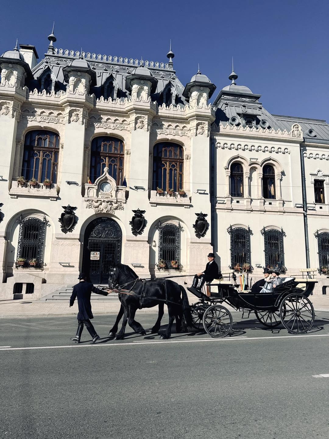 Horse-drawn carriage with a driver and two passengers in formal attire, pulled by a black horse in front of an ornate white stone building with arched windows and decorative details under a clear blue sky.