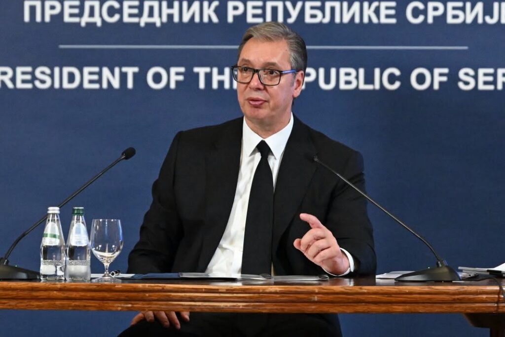 Man in a suit and glasses speaks at a press conference, seated behind a wooden table with microphones and water bottles. Blue backdrop reads'President of the Republic of Serbia'.