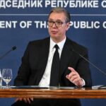 Man in a suit and glasses speaks at a press conference, seated behind a wooden table with microphones and water bottles. Blue backdrop reads 'President of the Republic of Serbia'.